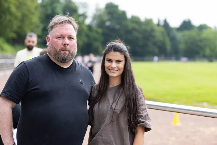 Ein Mann mit Bart und eine junge Frau lächeln in die Kamera. Im Hintergrund ist ein Sportplatz und weitere Personen zu sehen.