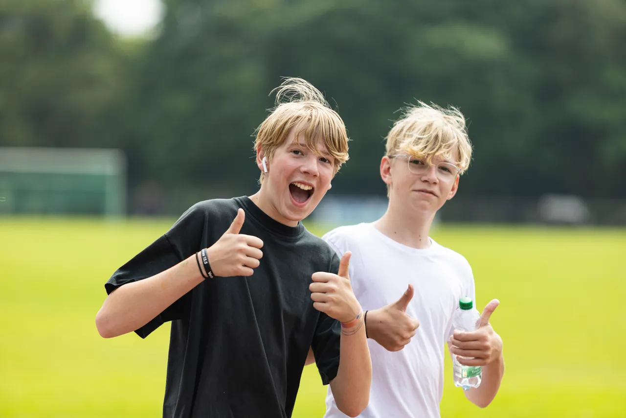 Zwei junge Männer mit blonden Haaren zeigen auf einem grünen Rasen Daumen hoch und lächeln in die Kamera. Im Hintergrund ist ein Sportplatz und Bäume zu sehen.