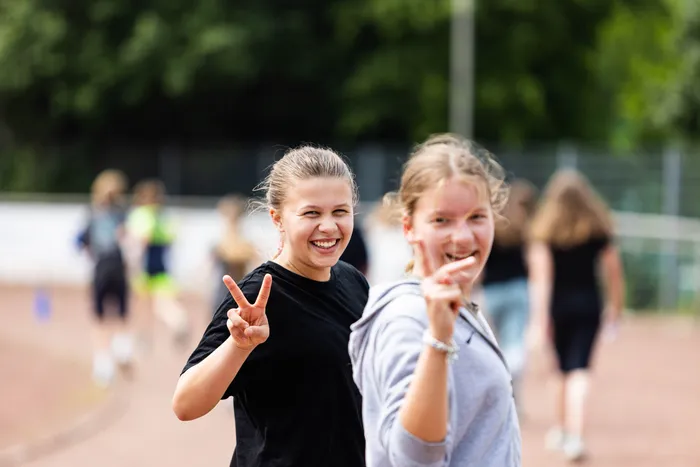 Zwei junge Frauen lächeln in die Kamera und zeigen beim Sponsorenlauf der Gesamtschule Kierspe Siegeszeichen. Im Hintergrund sind weitere Läufer auf einer roten Tartanbahn zu sehen.