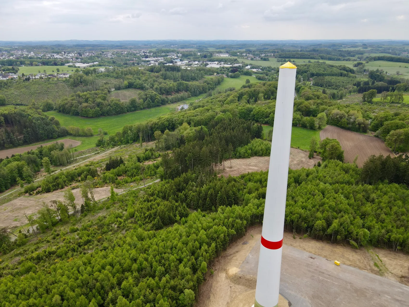 Weißer Turmschaft einer Windkraftanlage ragt inmitten grüner Landschaft und Wälder auf, im Hintergrund eine Stadt.