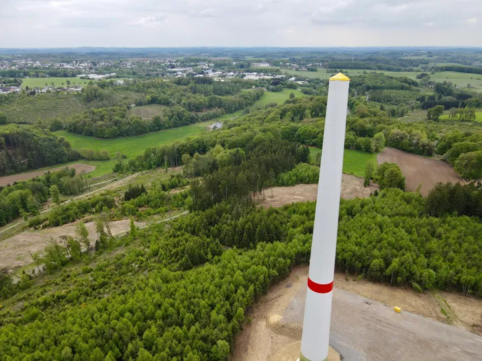Weißer Turmschaft einer Windkraftanlage ragt inmitten grüner Landschaft und Wälder auf, im Hintergrund eine Stadt.