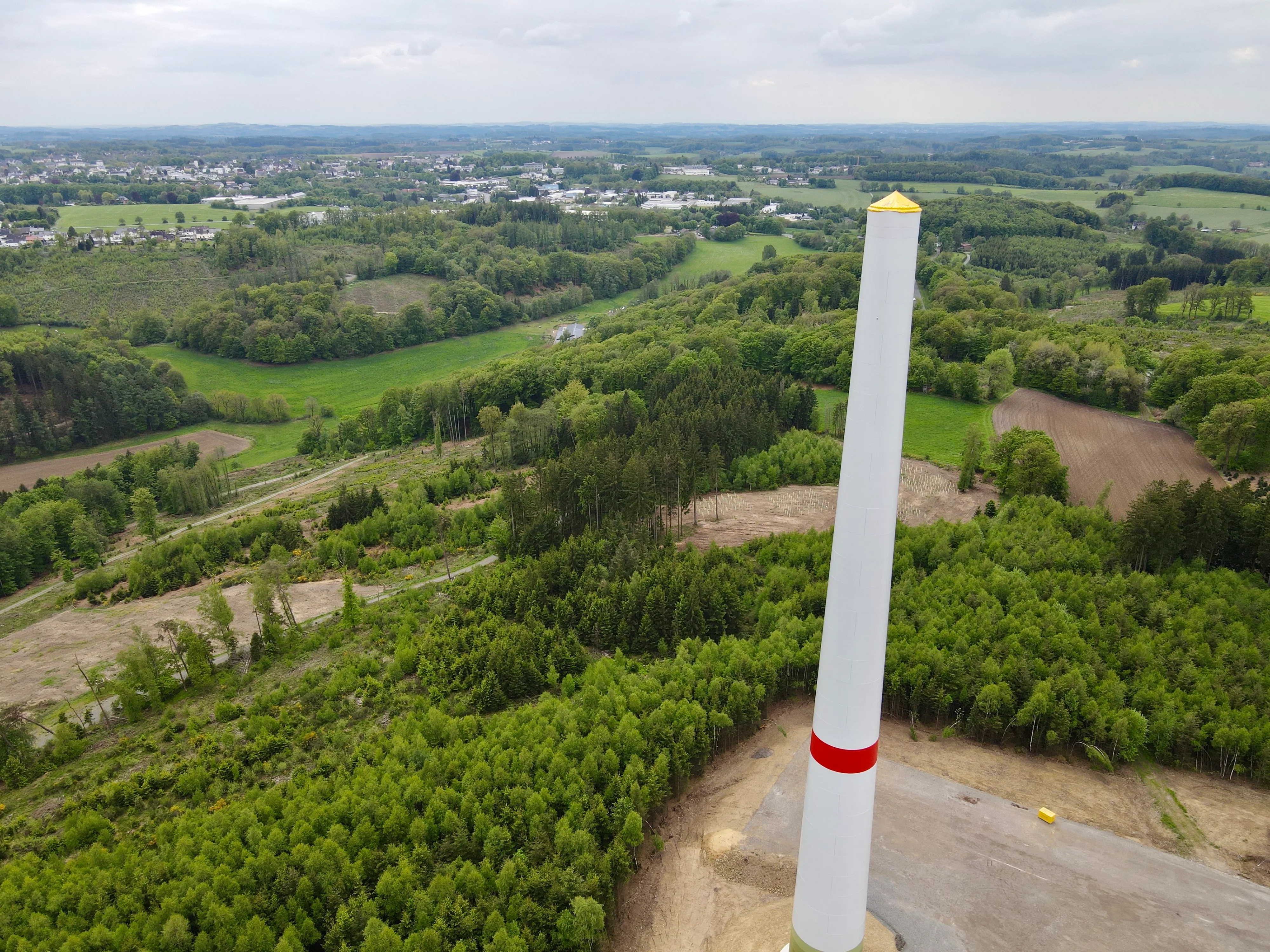 Weißer Turmschaft einer Windkraftanlage ragt inmitten grüner Landschaft und Wälder auf, im Hintergrund eine Stadt.