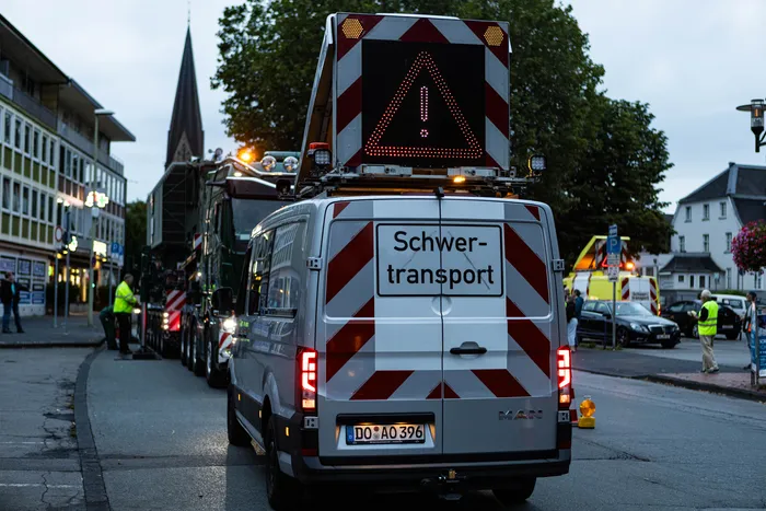 Ein weißer Transporter mit der Aufschrift „Schwer-transport“ und einem großen Warnschild auf dem Dach fährt vor einem Schwertransport.