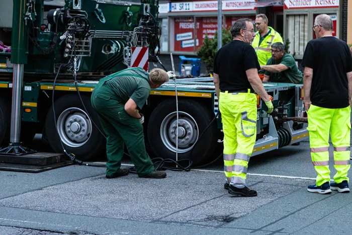 Männer in Arbeitskleidung und Warnwesten arbeiten an einem großen grünen Schwertransport-LKW.