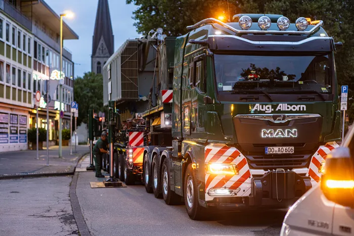 Ein grüner MAN-Schwertransport der Firma Aug. Alborn transportiert einen großen Transformator auf einer Stadtstraße bei Dämmerung.