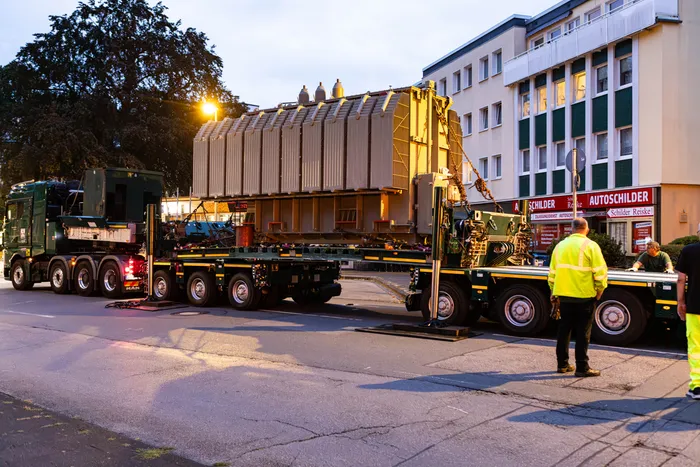 Ein großer, gelblicher Transformator wird auf einem Schwertransport über eine Straße befördert.