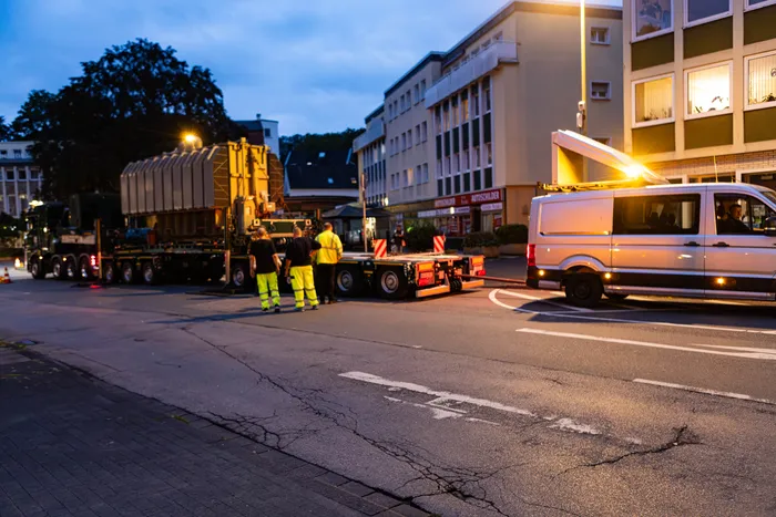 Schwertransport eines großen gelben Transformators auf einer Straße bei Dämmerung, daneben stehen Arbeiter in Warnwesten.