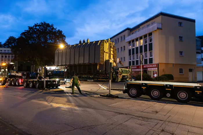 Ein großer Transformator wird auf einem mehrachsigen Schwertransport-Anhänger durch eine Straße bei Dämmerung transportiert.