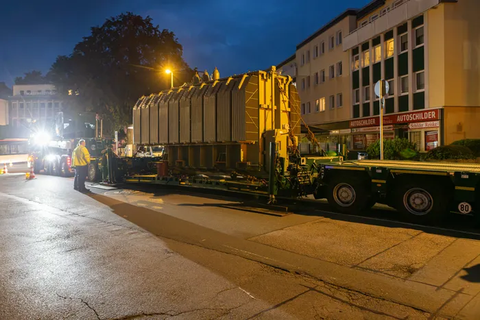 Ein großer Transformator auf einem Tieflader-Schwertransport wird nachts durch eine Stadtstraße befördert.