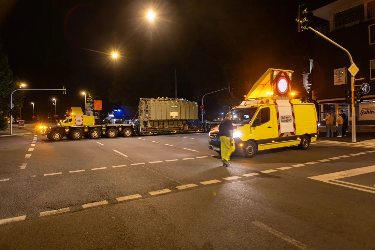 Gelber Schwertransport befördert einen großen grauen Transformator nachts über eine Straßenkreuzung.