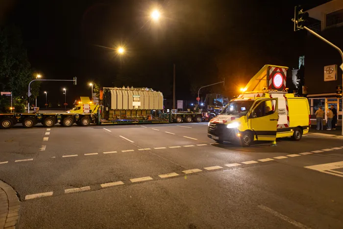 Ein gelbes Begleitfahrzeug fährt neben einem Schwertransport, der einen großen Transformator auf einer nächtlichen Stadtstraße befördert.