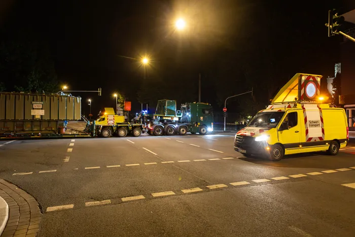 Zwei Schwertransporte mit großen Siemens-Containern auf einer nächtlich beleuchteten Straße, begleitet von einem gelben Begleitfahrzeug.