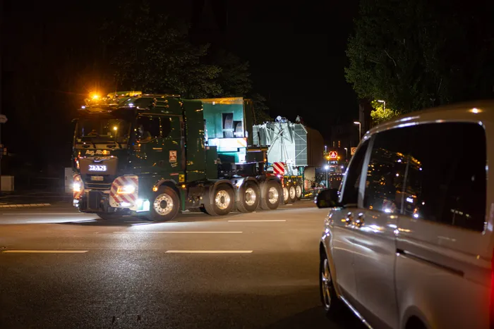 Ein grüner Schwertransport fährt nachts mit einem großen Transformator über eine Straße.