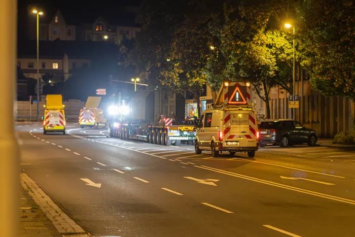 Zwei Schwertransporte fahren nachts auf einer Straße, darunter ein LKW mit schwerer Ladung und ein Begleitfahrzeug mit Warndreieck.