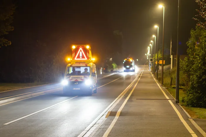 Ein Schwertransport mit leuchtenden Warnsignalen und einem großen dreieckigen Warnschild fährt nachts auf einer Straße.