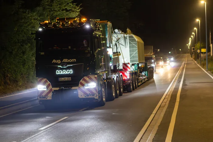 Ein MAN-Schwertransport transportiert nachts einen großen, weißen Transformator auf einer Straße.