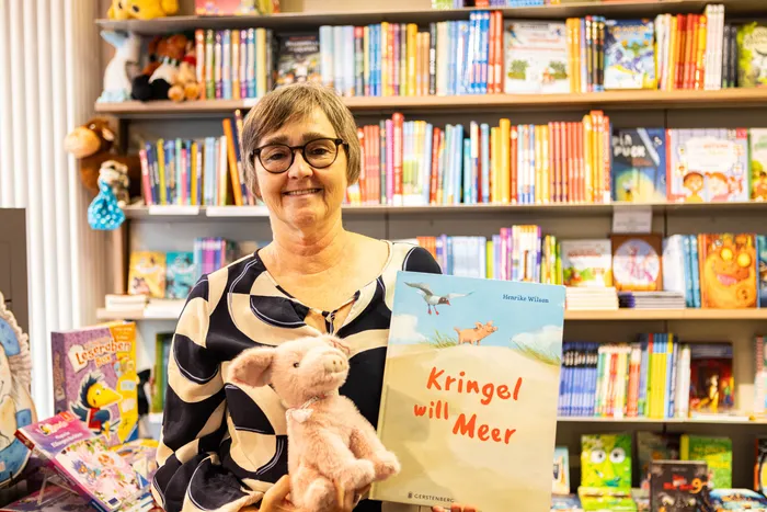 Kerstin Bestgen hält das Kinderbuch „Kringel will Meer“ und ein rosa Plüschtier vor einem Bücherregal.