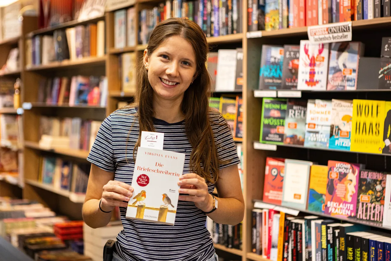 Lea Kempers hält in einer Buchhandlung das Buch „Die Briefeschreiberin“ von Virginia Evans mit einem „Mein Lesetipp“-Inlay hoch.