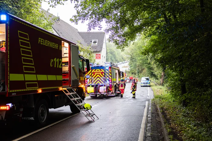 Ein Feuerwehrfahrzeug und Einsatzkräfte auf einer nassen Straße vor einem Haus am Waldrand.