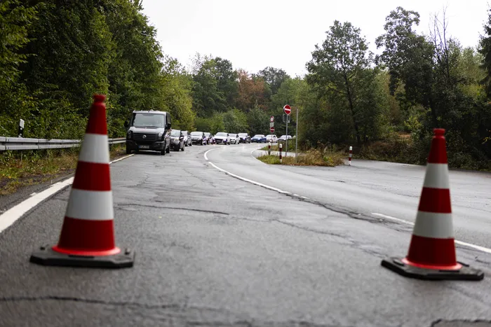Rot-weiße Leitkegel im Vordergrund einer nassen, kurvigen Straße, auf der sich im Hintergrund eine Reihe von Fahrzeugen durch eine bewaldete Gegend bewegt.