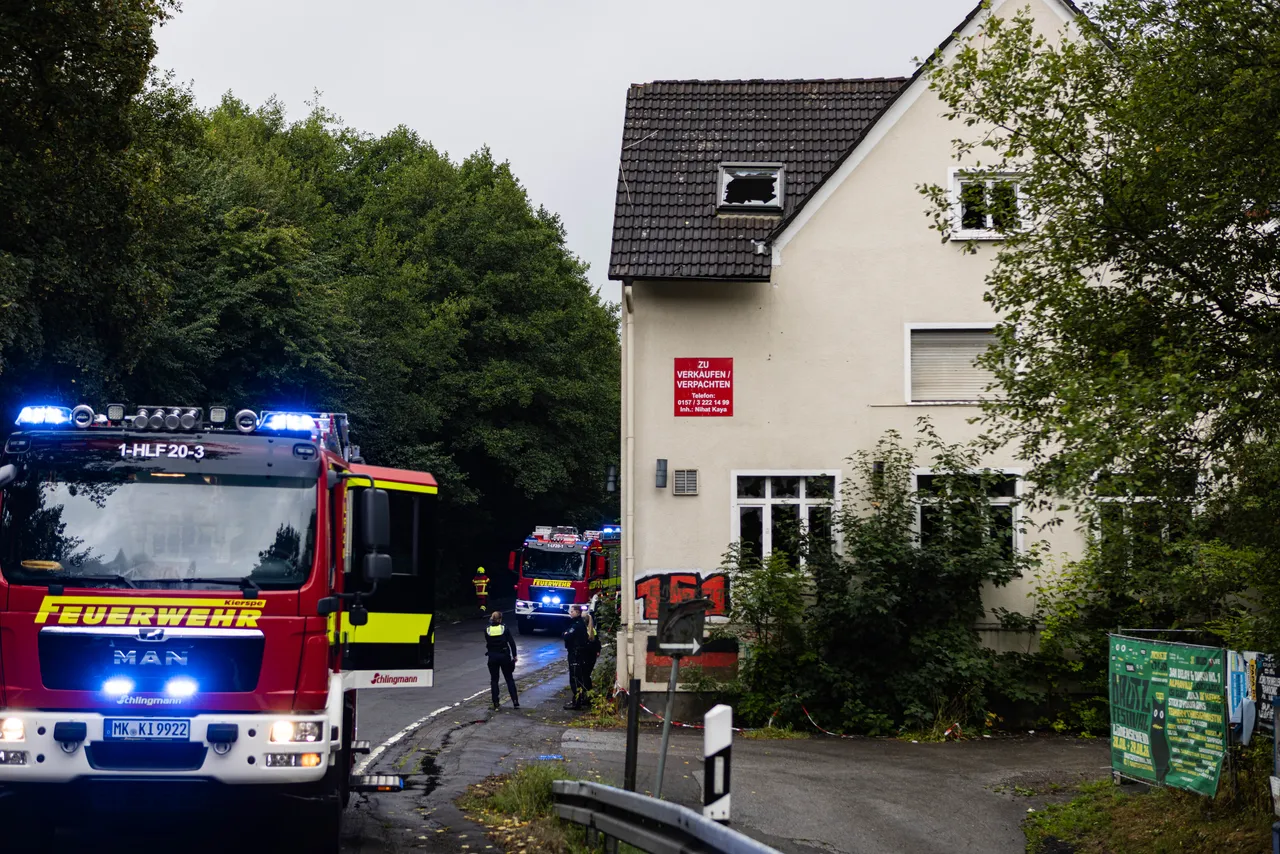 Ein Feuerwehrfahrzeug mit Blaulicht steht vor einem weißen Haus; im Hintergrund sind weitere Einsatzkräfte und ein weiteres Einsatzfahrzeug bei einem Brandeinsatz zu sehen.