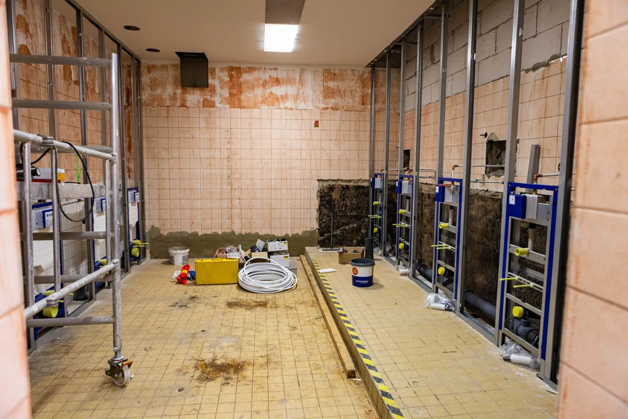 Baustelle in einem Schulgebäude mit Metallrahmen für Sanitärinstallationen, einem Gerüst und Baumaterialien auf dem gefliesten Boden.