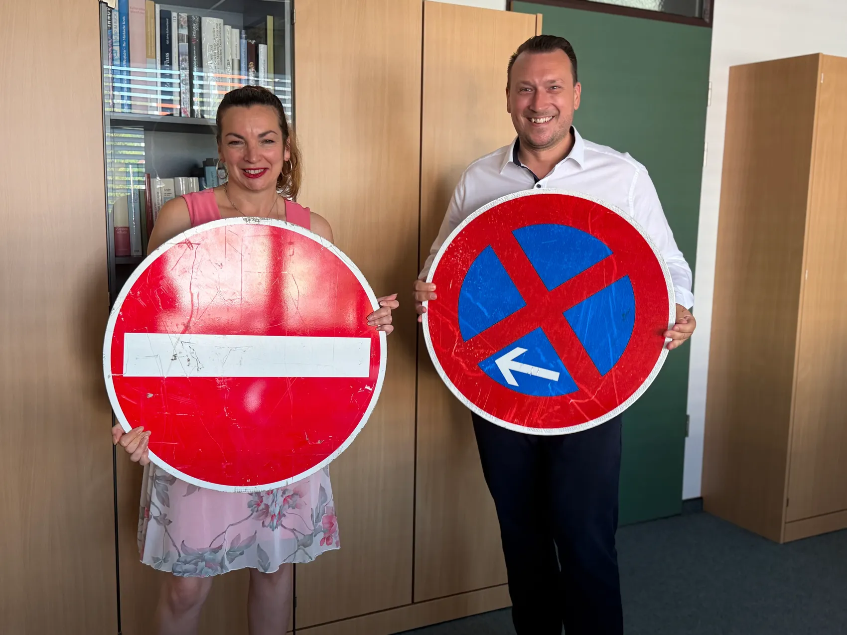 Zwei Personen halten jeweils ein Verkehrsschild: links ein rotes Schild mit weißem Balken für „Verbotene Einfahrt“, rechts ein rotes Schild mit blauem Kreuz und Pfeil für Parkregelung.