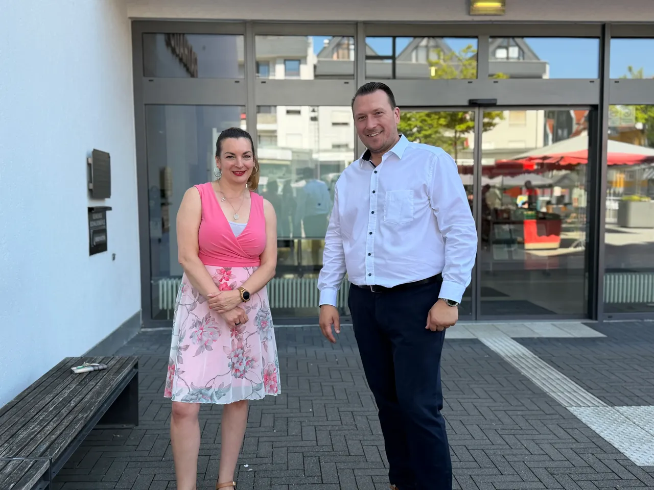 Zwei Personen, eine Frau in einem rosa Kleid und ein Mann in Hemd und Hose, stehen vor dem Eingang eines Gebäudes. Im Hintergrund sind weitere Personen und ein roter Sonnenschirm zu sehen.
