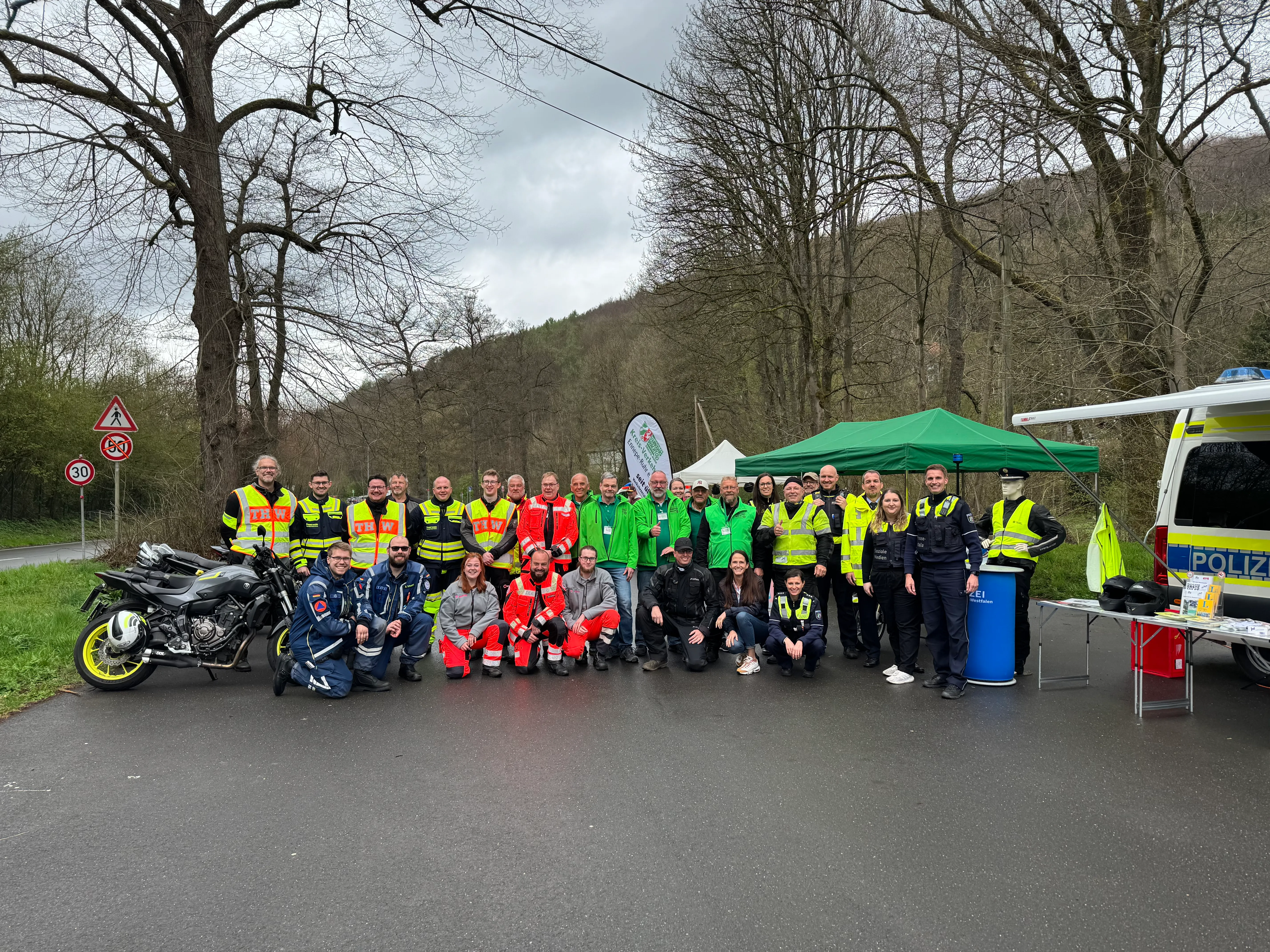 Gruppe von Einsatzkräften und Helfern in Warnwesten vor einem Zelt und einem Polizeiwagen am Straßenrand.