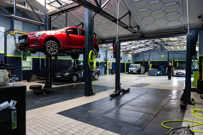 Ein roter Wagen ist in einer Autowerkstatt auf einer Hebebühne angehoben. Im Hintergrund stehen weitere Fahrzeuge und ein Mechaniker arbeitet.