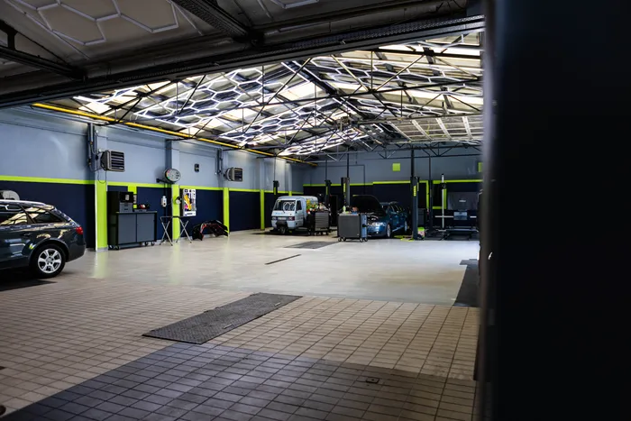 Eine hell erleuchtete Autowerkstatt mit mehreren Fahrzeugen und Hebebühnen. Im Hintergrund ist ein Schreibtisch mit einem Computer zu sehen.