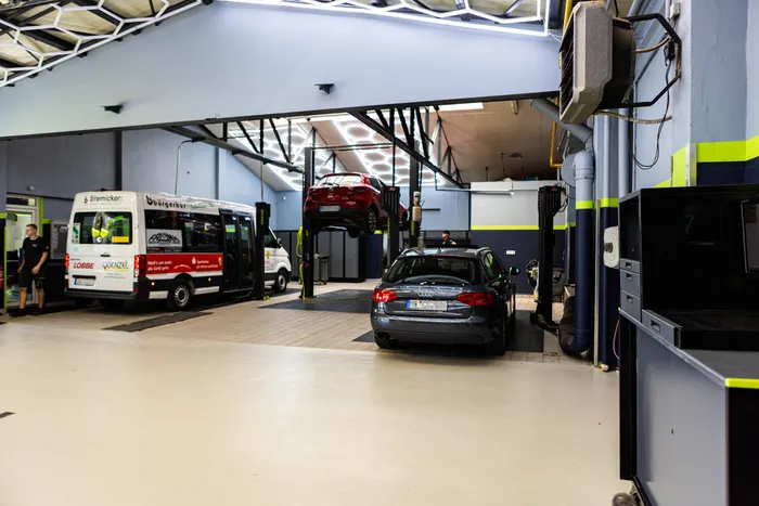 Ein rotes Auto steht auf einer Hebebühne in einer modernen, hellen Werkstatt. Im Hintergrund stehen zwei weiße Transporter und ein Mann in Arbeitskleidung.