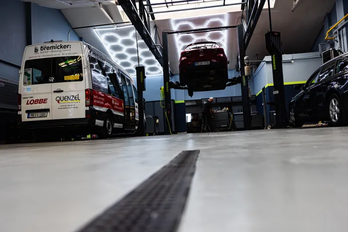 Ein roter Kleinwagen steht auf einer Hebebühne in einer modernen Autowerkstatt. Im Hintergrund ist ein roter Transporter mit der Aufschrift 