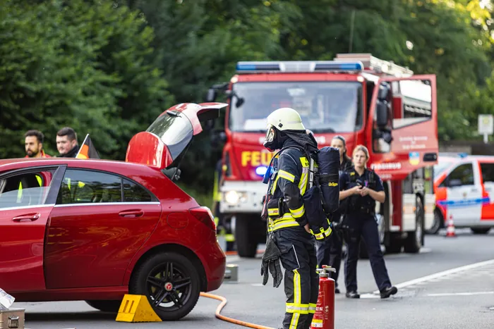 Feuerwehrleute stehen an einem Unfallort mit einem roten Auto und einem roten Feuerwehrauto im Hintergrund. Einsatzkräfte versorgen Verletzte nach einem Verkehrsunfall auf der Straße.