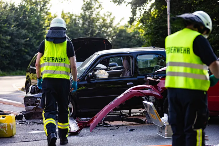 Zwei Feuerwehrleute in Warnwesten stehen vor einem stark beschädigten schwarzen Auto mit hochgebogener Motorhaube und Trümmerteilen auf der Straße. Im Hintergrund ist ein weiteres rotes Fahrzeug mit erheblichen Schäden zu erkennen.