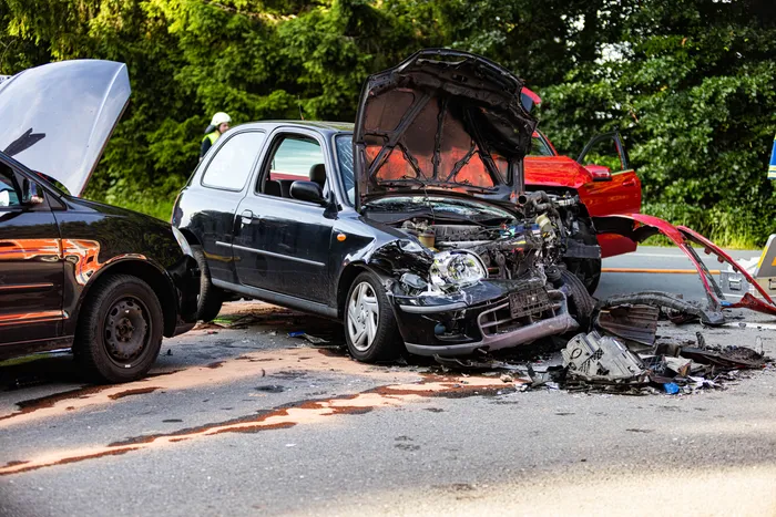 Schwer beschädigte schwarze Pkw mit geöffneter Motorhaube nach einem Verkehrsunfall auf einer Landstraße. Im Hintergrund ein rotes Fahrzeug und eine Person mit Helm. Öl und Trümmerteile liegen auf der Fahrbahn.