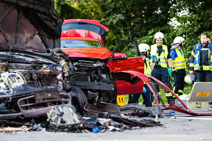Zwei stark beschädigte Autos nach einem Unfall auf einer Landstraße. Rettungskräfte in gelben Westen sind im Hintergrund zu sehen.