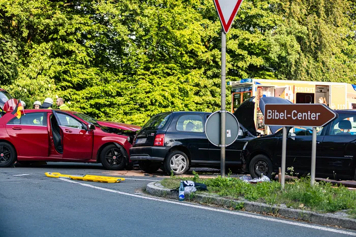 Mehrere beschädigte Autos blockieren eine Landstraße, umgeben von Einsatzfahrzeugen und Rettungskräften. Trümmerteile liegen auf der Fahrbahn.