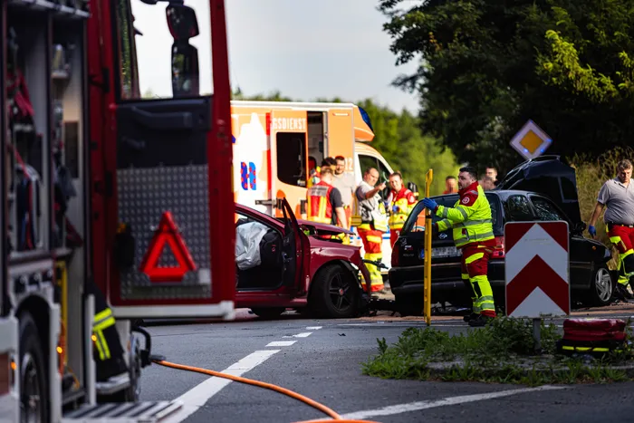 Rettungskräfte betreuen nach einem Unfall auf einer Landstraße mehrere beschädigte Fahrzeuge. Ein rotes Auto hat schwere Frontschäden, daneben steht ein Krankenwagen und ein Feuerwehrauto.