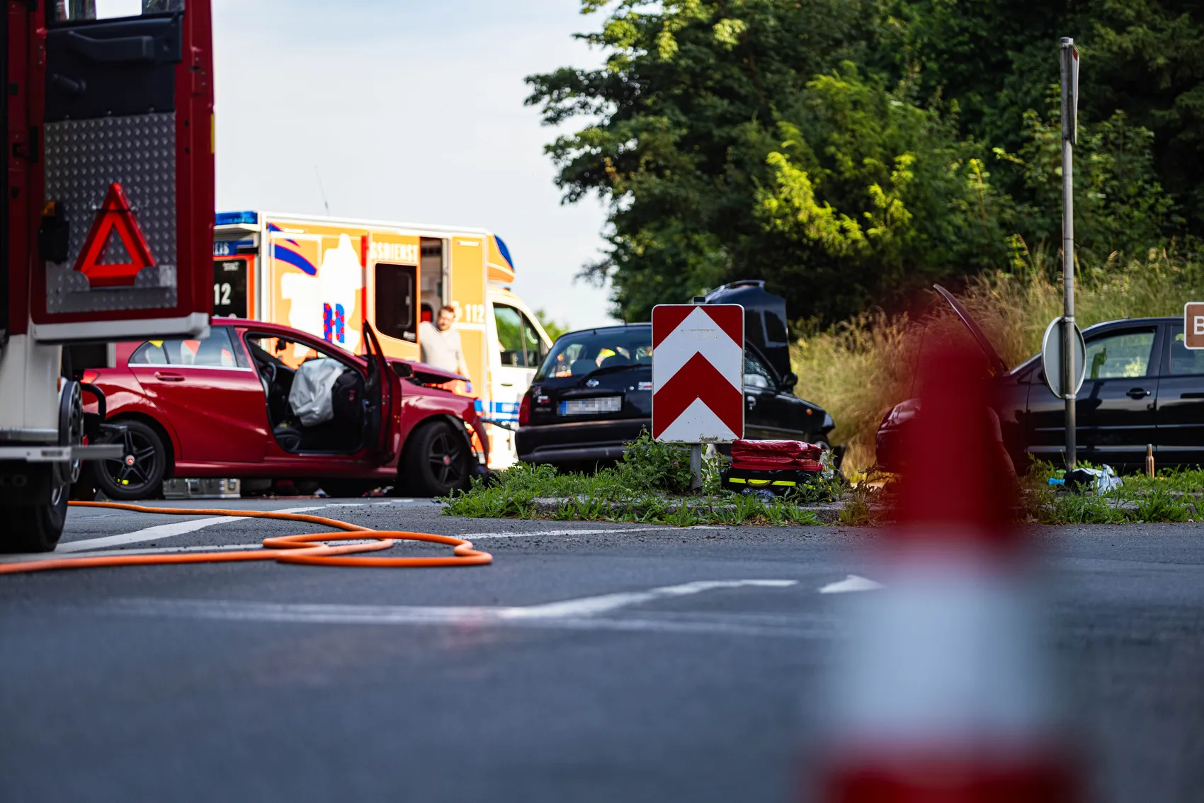 Ein rotes Auto mit erheblichen Schäden an der Fahrerseite steht auf einer Landstraße. Im Hintergrund sind ein Krankenwagen, ein Abschleppwagen und weitere Fahrzeuge zu sehen.