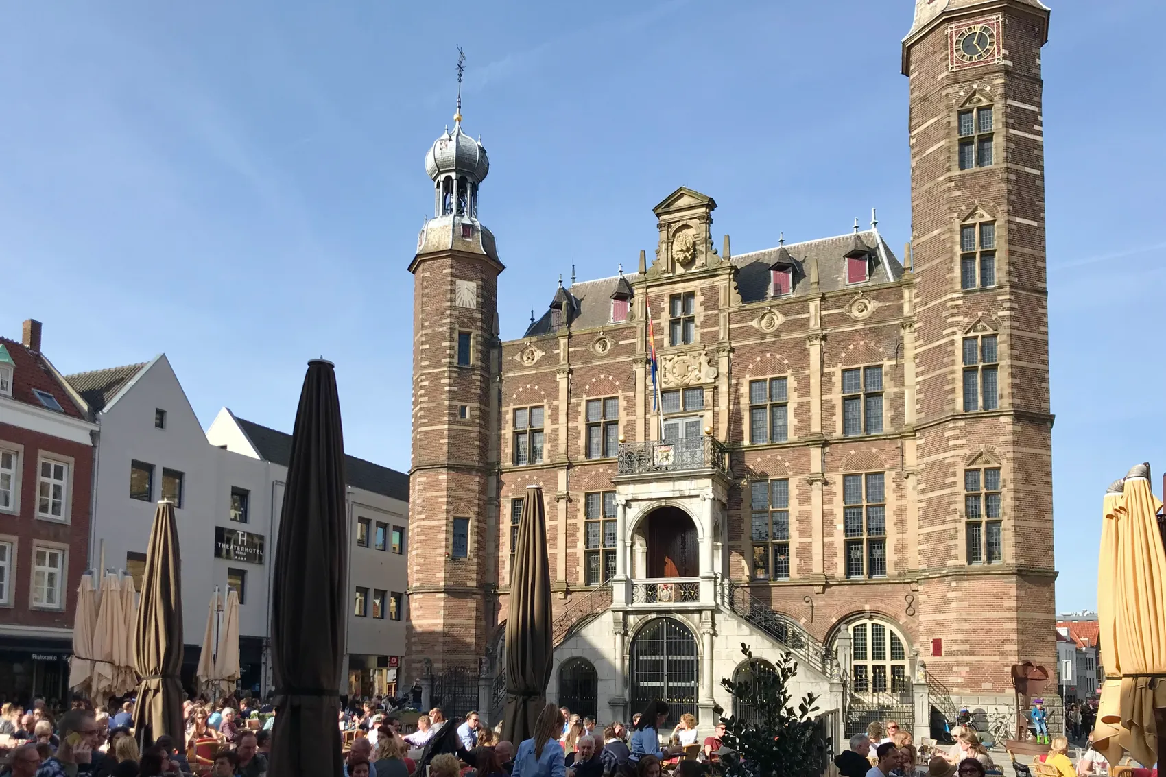 Das historische Rathaus von Venlo mit Ziergiebeln und einem Uhrturm erhebt sich über einem belebten Platz mit Sonnenschirmen und Menschen.