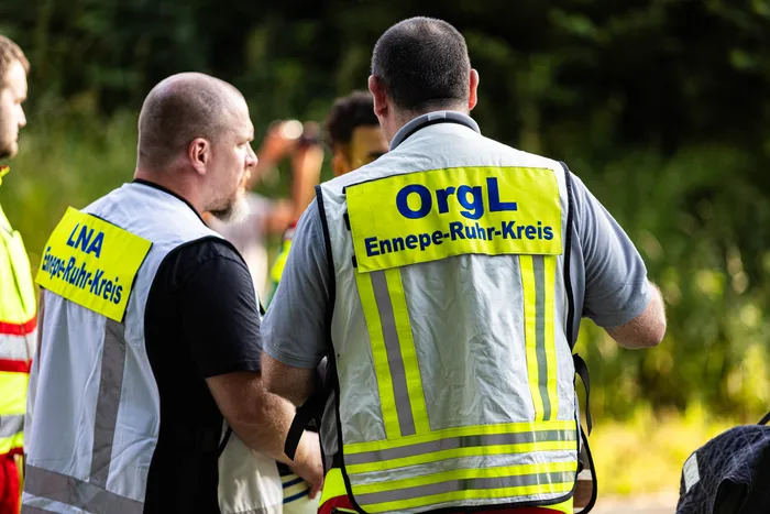 Zwei Einsatzkräfte mit Warnwesten „OrgL Ennepe-Ruhr-Kreis“ und „LNA Ennepe-Ruhr-Kreis“ stehen mit dem Rücken zum Betrachter da. Im Hintergrund sind weitere Personen und Bäume zu sehen.