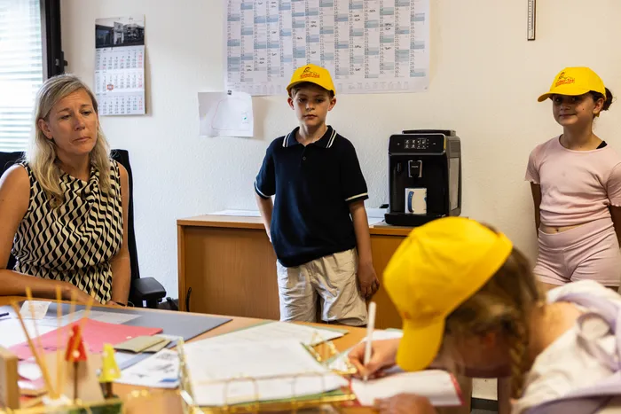 Eine Frau und drei Kinder stehen in einem Büro. Die Kinder tragen gelbe Hüte und zwei von ihnen schauen auf ein Dokument, das auf einem Tisch liegt. Im Hintergrund hängt ein Kalender.