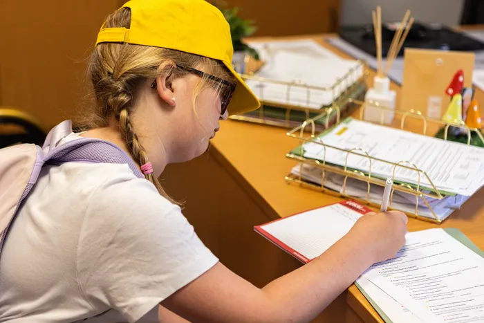 Ein Mädchen mit gelber Kappe und Sonnenbrille schreibt Notizen auf ein Formular. Im Hintergrund stehen Büroartikel und Unterlagen auf einem Schreibtisch.