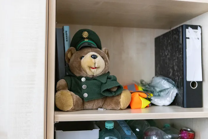 Ein brauner Teddybär mit grüner Uniform und einer Schirmmütze sitzt in einem Regal zwischen Aktenordnern, Büchern und anderen Gegenständen. Im Vordergrund stehen Flaschen.