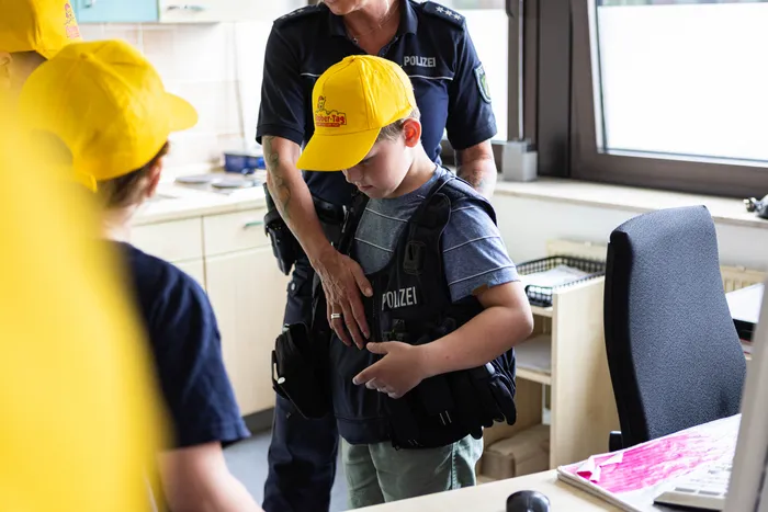 Ein Polizist hilft einem Jungen beim Anlegen einer Schutzweste. Der Junge trägt eine gelbe Kappe mit der Aufschrift 
