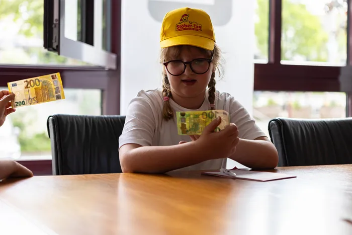 Ein Mädchen mit Zöpfen und Brille sitzt an einem Tisch und hält Geldscheine in der Hand. Im Hintergrund ist ein Fenster und ein weiteres Mädchen mit einem 200-Euro-Schein zu sehen.