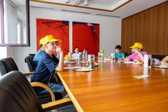 Kinder sitzen an einem großen Konferenztisch und essen. Ein Junge mit gelber Kappe blickt nachdenklich in die Ferne, während andere Kinder im Hintergrund zu sehen sind. Auf dem Tisch stehen Getränke und Snacks.