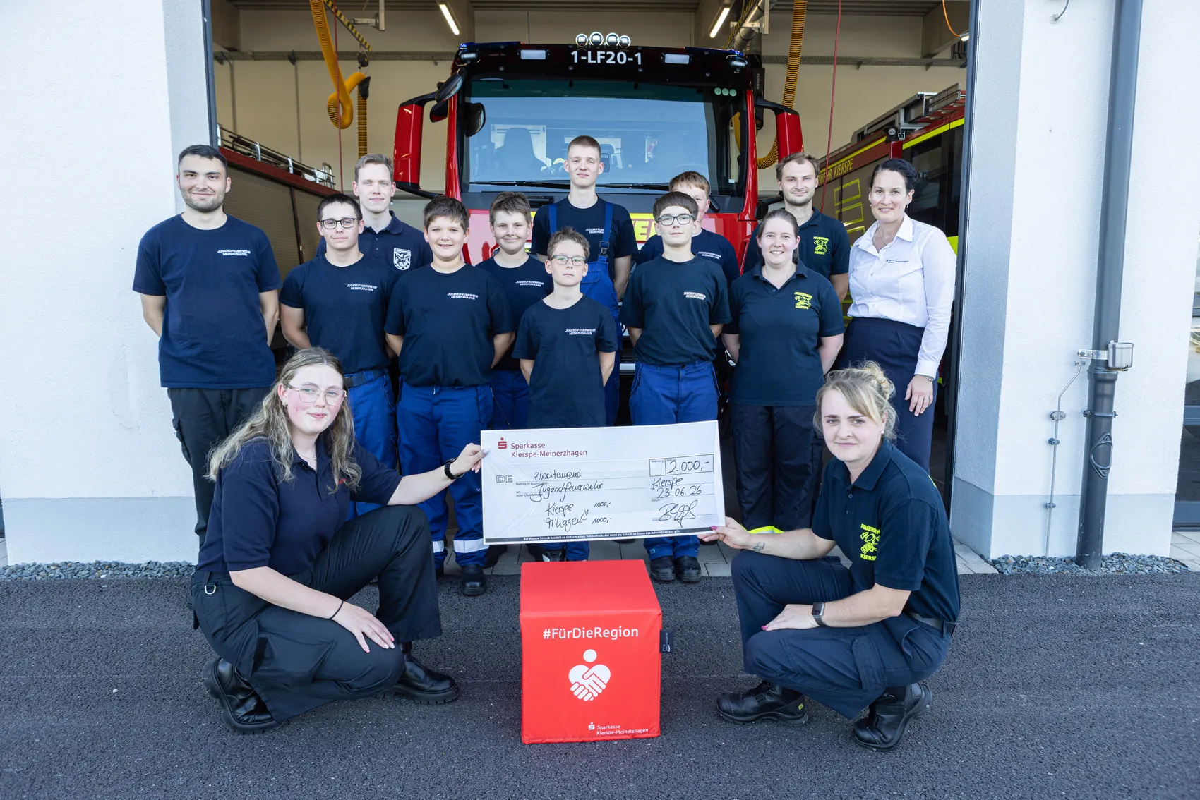 Gruppe von Feuerwehrleuten, darunter Jugendliche, posieren vor einem Feuerwehrauto. Zwei Frauen halten einen übergroßen Scheck der Sparkasse Kierspe-Meinerzhagen entgegen.