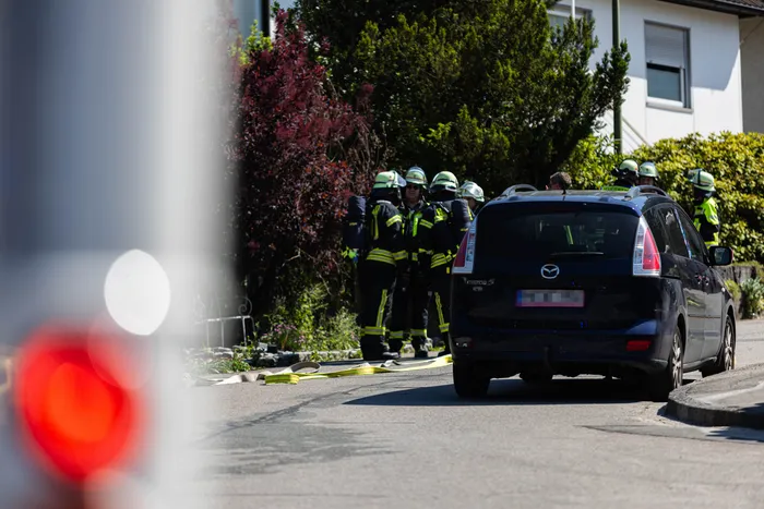 Feuerwehrleute in gelben Schutzanzügen und Helmen sind an einem dunklen Kombi im Einsatz, während ein Schlauch auf der Straße liegt. Im Hintergrund steht ein Wohnhaus mit Büschen und Bäumen.